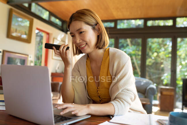 Smiling asian woman using laptop and talking on smartphone, working from home. at home in isolation during quarantine lockdown. — Stock Photo
