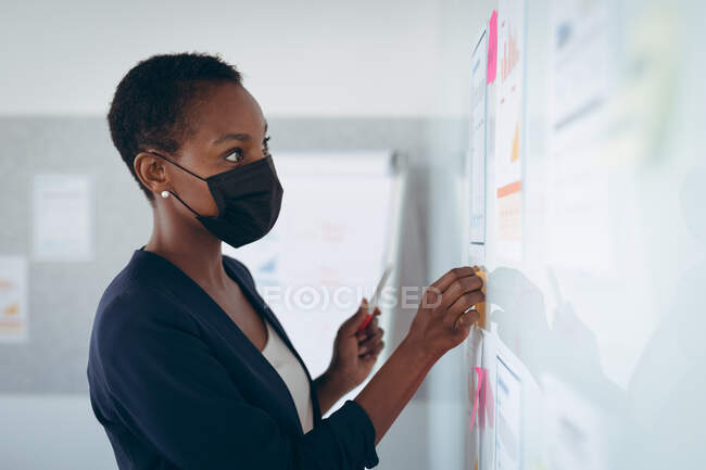 Empresaria afroamericana con máscara facial tomando notas y agregando post-ins en la pared. negocio creativo independiente en una oficina moderna durante coronavirus covid 19 pandemia. - foto de stock