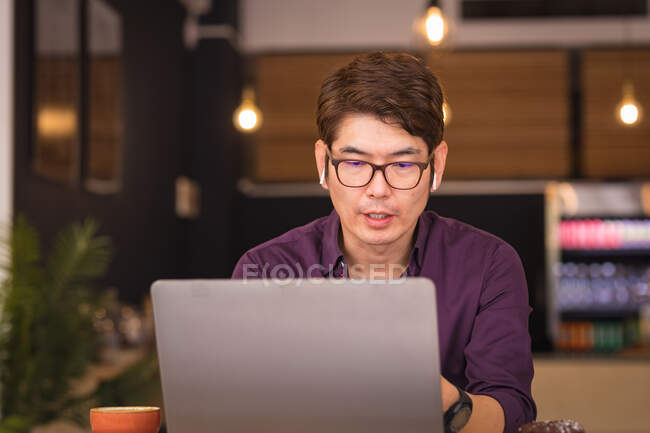 Asian businessman using laptop and wireless earphones in cafe. business travel, digital nomad on the go out and about in city concept. — Stock Photo