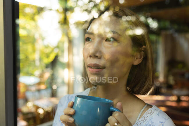 Retrato de mulher asiática sorridente segurando caneca de chá e olhando pela janela. em casa em isolamento durante o confinamento de quarentena. — Fotografia de Stock