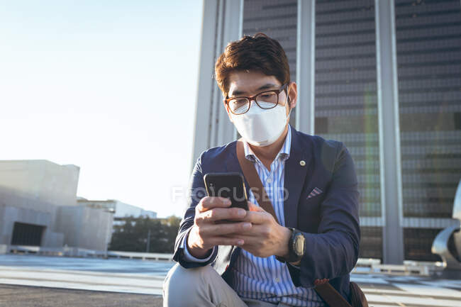 Asian businessman wearing face mask using smartphone in city street. digital nomad out and about in city during covid 19 pandemic concept. — Stock Photo