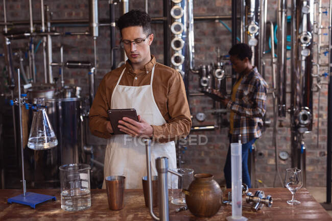 Кавказький робітник одягнений в фартух, використовуючи цифрову табличку на джинській винокурні. Виробництво алкоголю і фільтрація. — стокове фото