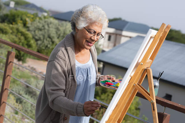 Gemischte Rennen Seniorin Malerei auf Leinwand auf dem Balkon. Isolationshaft während der Quarantäne. — Stockfoto