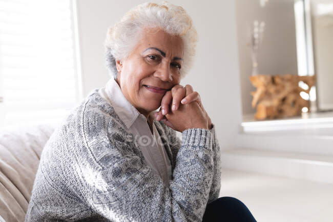 Портрет старшої жінки змішаної раси, яка сидить на дивані, дивлячись на камеру і посміхаючись. Перебуваючи вдома в ізоляції під час карантину.. — стокове фото