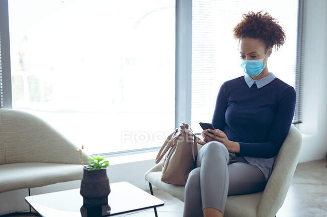 Деловая женщина смешанной расы в маске и со смартфоном. работа в современном офисе во время пандемии коронавируса ковида 19. — стоковое фото