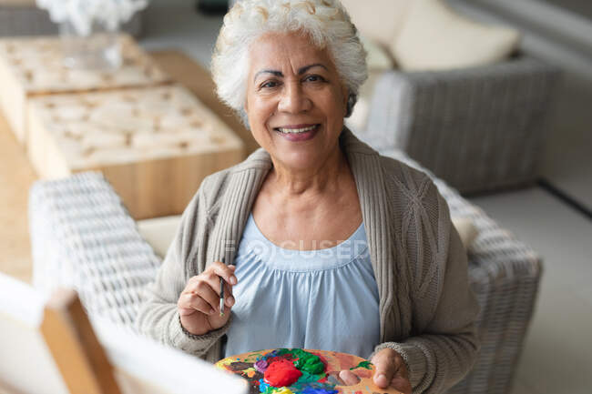 Portrait of mixed race senior woman painting on canvas in living room. staying at home in isolation during quarantine lockdown. — Stock Photo