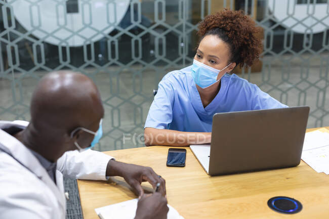 Diverso médico masculino y enfermera con máscaras faciales, el uso de la computadora portátil y hablar. medicina, salud y servicios de salud durante la pandemia del coronavirus covid 19. - foto de stock