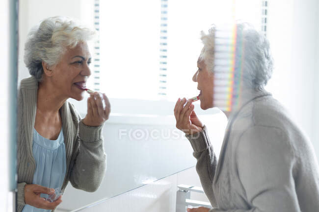 Mujer mayor de raza mixta mirando su reflejo en el espejo con lápiz labial. permanecer en casa aislado durante el bloqueo de cuarentena. - foto de stock