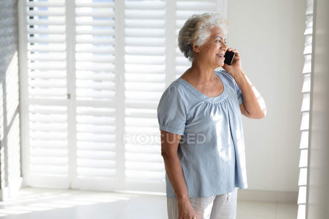 Mulher sênior de raça mista falando no smartphone ao lado da janela. ficar em casa em isolamento durante o confinamento de quarentena. — Fotografia de Stock