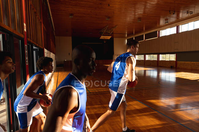 Equipo de baloncesto masculino diverso sosteniendo pelotas y entrando al gimnasio. baloncesto, entrenamiento deportivo en una cancha cubierta. - foto de stock
