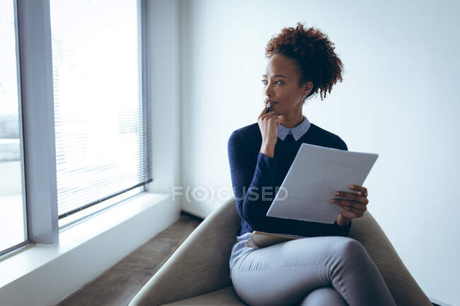 Empresaria mixta pensando, sentada al lado de la ventana y sosteniendo documentos. trabajar en un negocio creativo independiente. - foto de stock