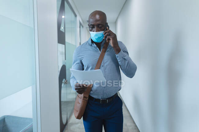 African american businessman wearing face mask and talking on smartphone. work at a modern office during covid 19 coronavirus pandemic. — Stock Photo