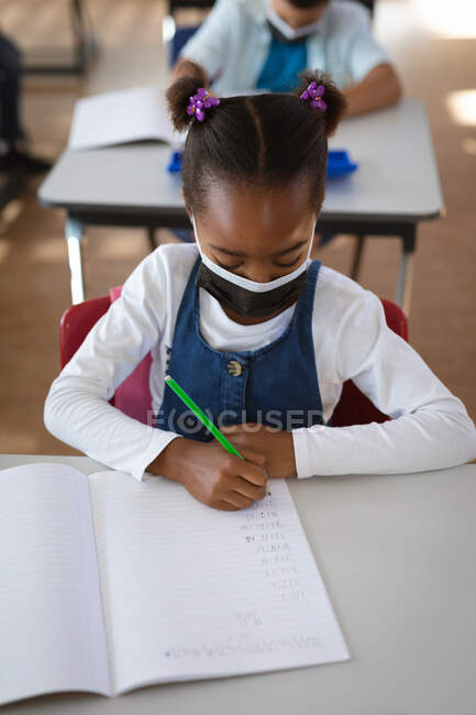 Ragazza afroamericana indossando maschera viso studiare mentre seduto sulla scrivania in classe a scuola. igiene e distanza sociale a scuola durante la covd 19 pandemia — Foto stock