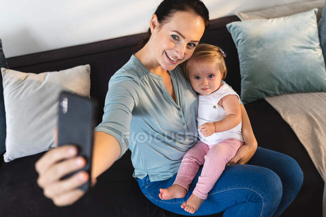 Kaukasische Mutter lächelt, während sie ein Selfie mit ihrem Baby macht, das zu Hause auf der Couch sitzt. Mutterschaft, Liebe und Babypflege-Konzept — Stockfoto