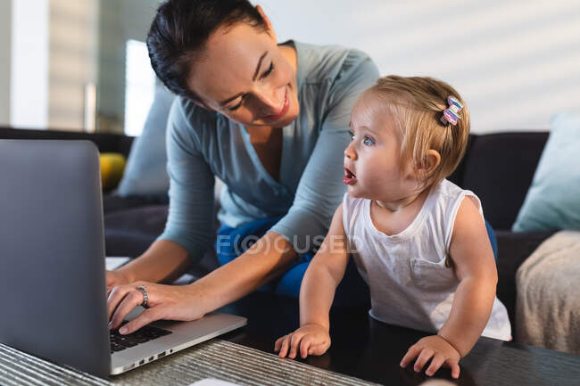 Sonriente madre caucásica mirando a su bebé usando el portátil mientras trabaja desde casa. maternidad, amor y cuidado del bebé - foto de stock