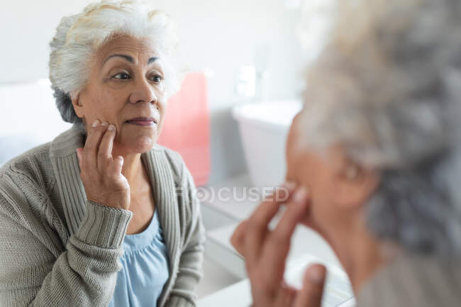 Старшая расовая женщина, смотрящая на свое отражение в зеркале. оставаться дома в изоляции во время карантинной изоляции. — стоковое фото