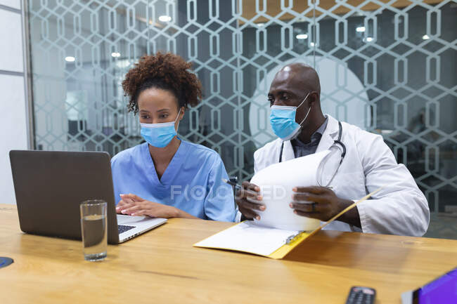 Médico masculino diverso e enfermeira feminina usando máscaras faciais, usando laptop e conversando. medicina, saúde e serviços de saúde durante coronavírus covid 19 pandemia. — Fotografia de Stock