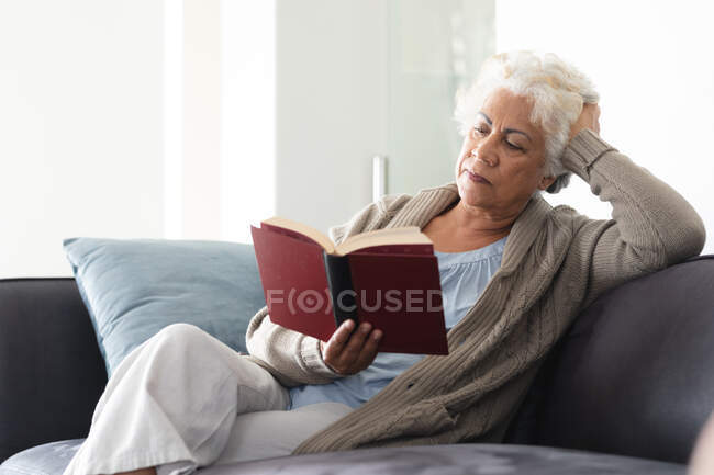Mulher sênior de raça mista sentada no livro de leitura do sofá. ficar em casa em isolamento durante o confinamento de quarentena. — Fotografia de Stock