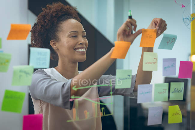 Femme d'affaires mixte prenant des notes sur un panneau de verre et souriant. travailler dans une entreprise créative indépendante. — Photo de stock