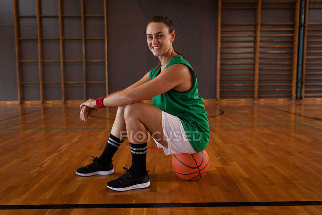 Portrait of caucasian female basketball player sitting on ball. basketball, sports training at an indoor court. — Stock Photo