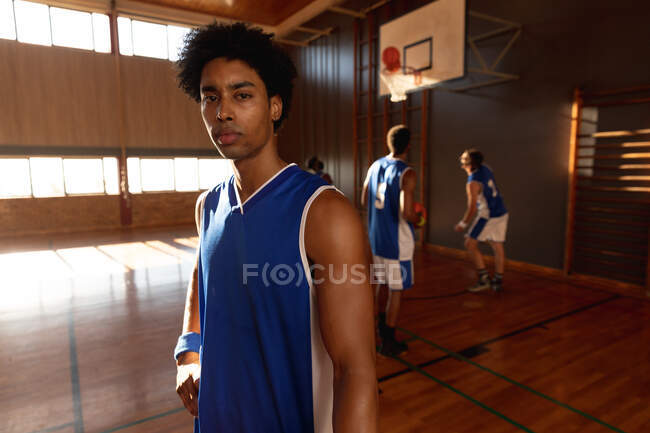 Retrato de mestiço jogador de basquete masculino com equipe em segundo plano. basquete, treinamento esportivo em uma quadra interna. — Fotografia de Stock
