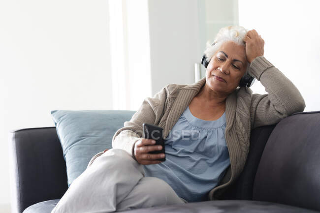 Mulher sênior de raça mista sentada no sofá usando fones de ouvido e usando smartphone. ficar em casa em isolamento durante o confinamento de quarentena. — Fotografia de Stock