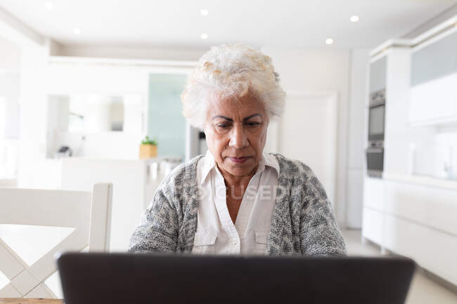 Mujer mayor de raza mixta sentada en la mesa usando un portátil. permanecer en casa aislado durante el bloqueo de cuarentena. - foto de stock