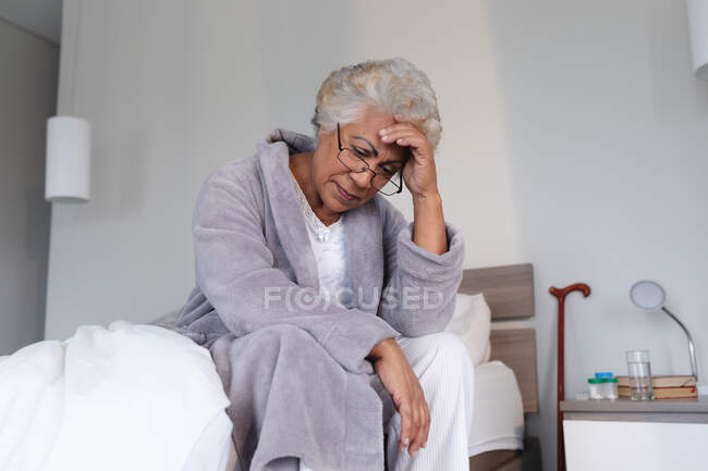 Mulher sênior de raça mista sentada na cama segurando a cabeça em pensamento. ficar em casa em isolamento durante o confinamento de quarentena. — Fotografia de Stock