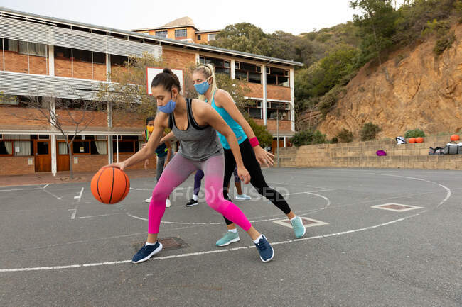 Equipe de basquete feminina diversa usando máscaras faciais e praticar driblando bola. treinamento de basquete em uma quadra urbana ao ar livre durante coronavírus covid 19 pandemia. — Fotografia de Stock