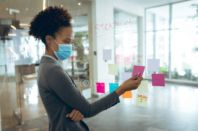Gemischte Rasse Geschäftsfrau mit Mundschutz und Notizen auf Glasplatte. Arbeit in einem modernen Büro während der Covid 19 Coronavirus-Pandemie. — Stockfoto