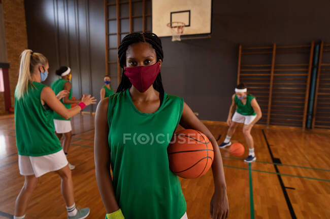 Retrato de una jugadora de baloncesto afroamericana usando máscara facial con equipo en el fondo. baloncesto, entrenamiento deportivo en una cancha de interior durante la pandemia de coronavirus covid 19. - foto de stock