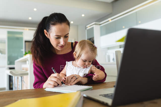 Mãe caucasiana segurando seu bebê tomando notas enquanto trabalhava em casa. maternidade, amor e conceito de cuidado do bebê — Fotografia de Stock