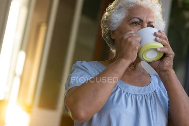 Старшая женщина смешанной расы пьет кофе рядом с дверью. оставаться дома в изоляции во время карантинной изоляции. — стоковое фото