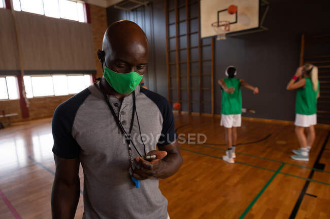 Entraîneur afro-américain de basket-ball portant un masque facial avec une équipe en arrière-plan. basket-ball, entraînement sportif sur un terrain intérieur pendant une pandémie de coronavirus covid 19. — Photo de stock