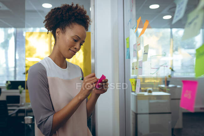 Деловая женщина смешанной расы делает записи на стеклянной доске и улыбается. работа в независимом креативном бизнесе. — стоковое фото