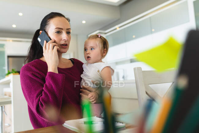 Кавказская мать держит своего ребенка, разговаривая по смартфону, работая из дома. материнство, любовь и уход за ребенком — стоковое фото