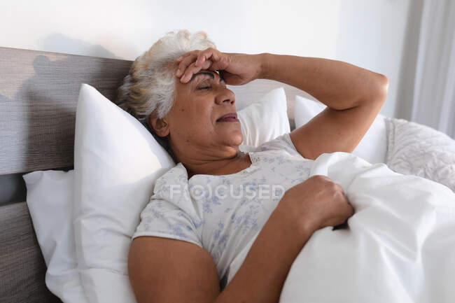 Mulher sênior de raça mista deitada na cama segurando a cabeça em pensamento. ficar em casa em isolamento durante o confinamento de quarentena. — Fotografia de Stock