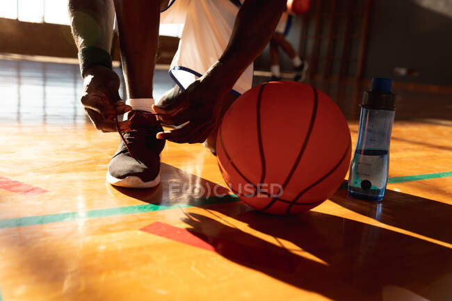 Jugador de baloncesto masculino atando zapatos con bola y agua. baloncesto, entrenamiento deportivo en una cancha cubierta. - foto de stock