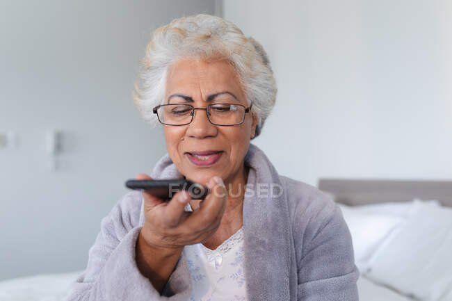 Mulher sênior de raça mista sentada na cama falando no smartphone. ficar em casa em isolamento durante o confinamento de quarentena. — Fotografia de Stock