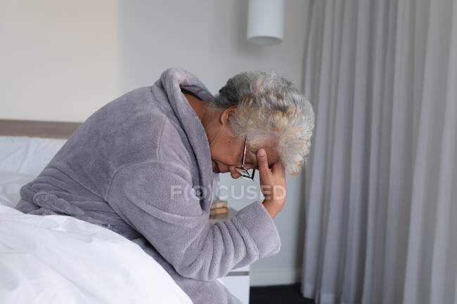Змішана раса старша жінка сидить на ліжку, тримаючи голову в думці. перебування вдома в ізоляції під час карантину . — стокове фото