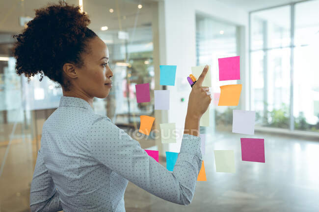 Gemischte Geschäftsfrau, die Notizen auf Glastafeln macht und denkt. Arbeit in einem unabhängigen kreativen Unternehmen. — Stockfoto