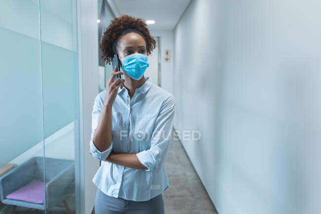 Femme d'affaires mixte portant un masque facial et parlant sur smartphone. travailler dans un bureau moderne pendant la pandémie de coronavirus covid 19. — Stock Photo