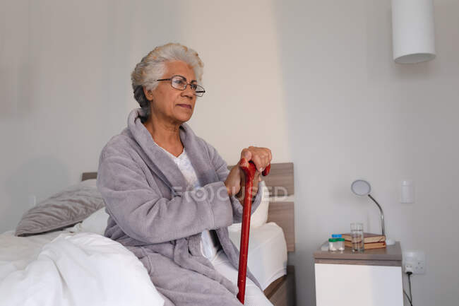 Змішана раса старша жінка сидить на ліжку тримаючи тростину. Перебуваючи вдома в ізоляції під час карантину.. — стокове фото