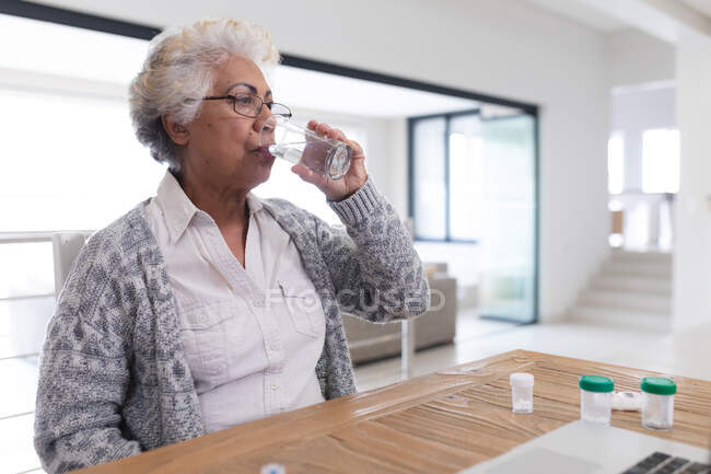 Mulher idosa de raça mista sentada à mesa a tomar medicamentos. ficar em casa em isolamento durante o confinamento de quarentena. — Fotografia de Stock