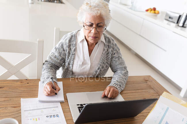 Mulher sênior de raça mista sentada à mesa usando laptop. ficar em casa em isolamento durante o confinamento de quarentena. — Fotografia de Stock