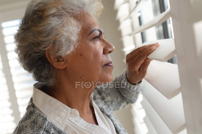 Mulher sénior de raça mista a olhar pela janela. ficar em casa em isolamento durante o confinamento de quarentena. — Fotografia de Stock
