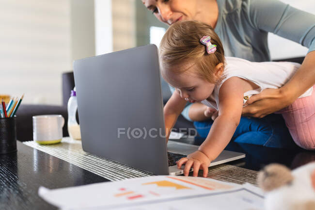 Madre caucásica sosteniendo a su bebé mientras intenta tocar el portátil en casa. maternidad, amor y cuidado del bebé - foto de stock