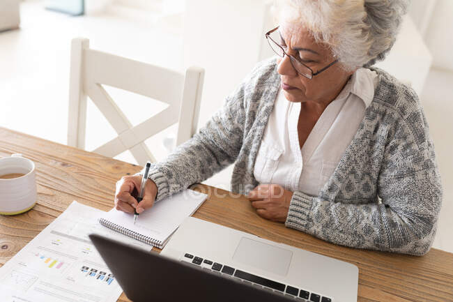 Eine gemischte Seniorin sitzt am Tisch und macht sich Notizen. Isolationshaft während der Quarantäne. — Stockfoto