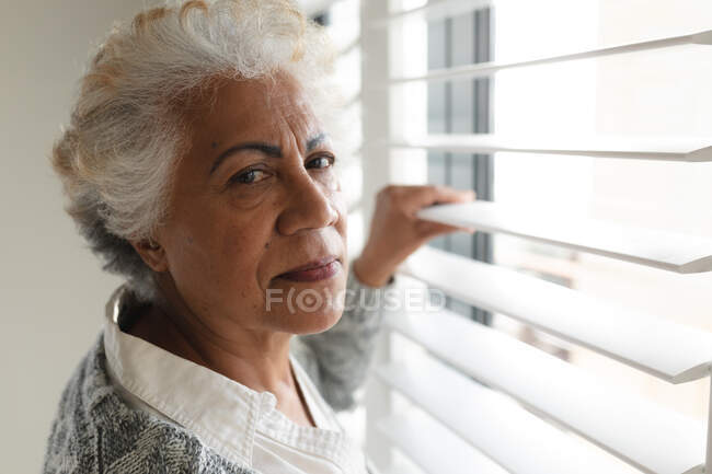 Porträt einer Seniorin mit gemischter Rasse, die in die Kamera am Fenster blickt. Isolationshaft während der Quarantäne. — Stockfoto