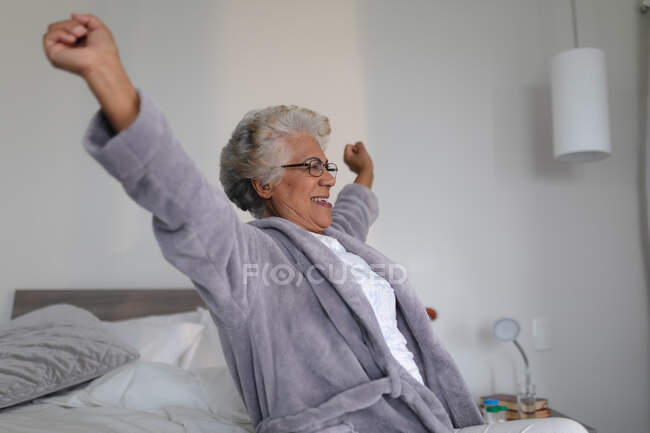 Mujer mayor de raza mixta sentada en la cama ensanchando sus brazos. permanecer en casa aislado durante el bloqueo de cuarentena. - foto de stock